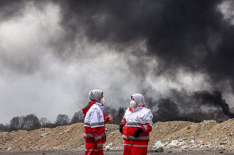Des femmes membres de la société du Croissant-Rouge iranien se tiennent près des panaches de fumée provenant d'un incendie suite à une frappe aérienne nocturne sur la raffinerie de pétrole de Shahran, dans le nord-ouest de Téhéran, en Iran, le 8 mars 2026. /VCG