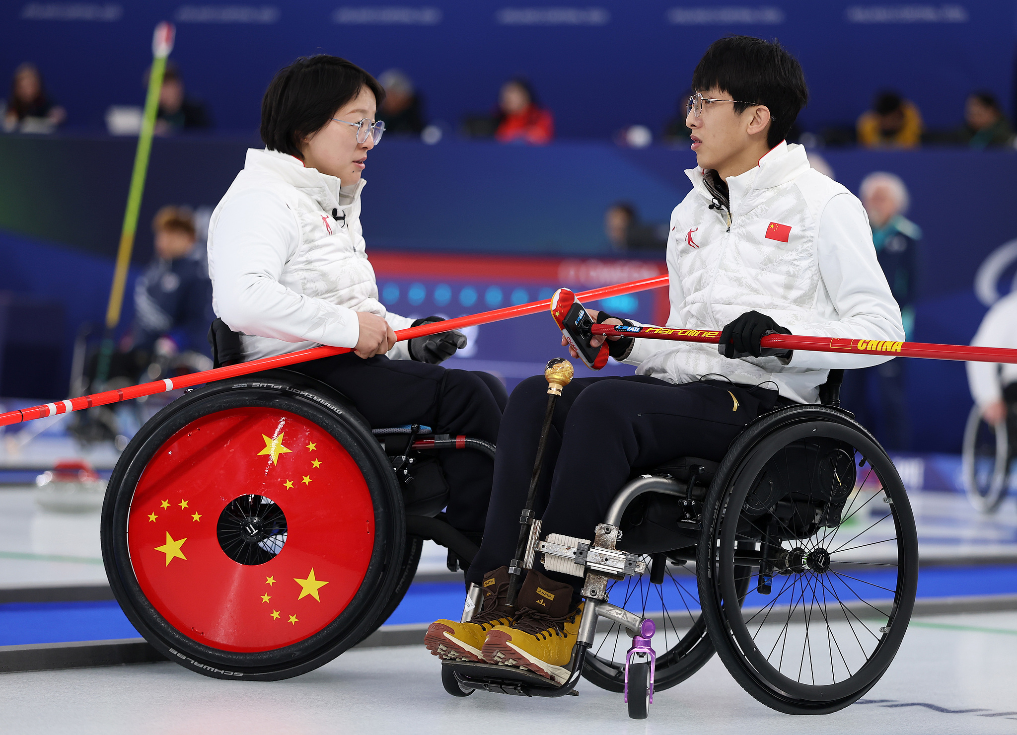 Wang Meng (à gauche) et Yang Jinqiao, de Chine, discutent de stratégie lors d'un premier tournoi à la ronde en double mixte de curling en fauteuil roulant contre l'Italie aux Jeux paralympiques d'hiver de Milan Cortina 2026 à Cortina d'Ampezzo, en Italie, le 5 mars 2026. /VCG
