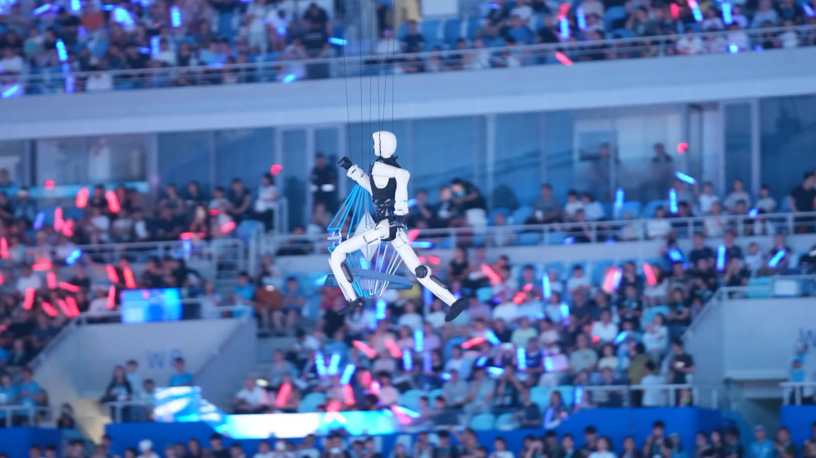 Un robot humanoïde est soulevé au-dessus de l'arène lors des Jeux mondiaux du robot humanoïde 2026, avec des spectateurs en arrière-plan, le 25 août 2026. Qu Bo/CGTN